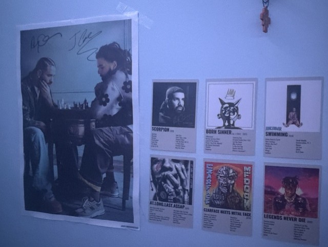 A poster displaying the artists Drake and J. Cole in a still from the music video for "First Person Shooter Mode", a song that became popular at parties and other events. Listening to music with others at parties is a popular way to connect, whether it's singing along to the lyrics with friends, striking up a conversation over a shared interest in the song, or simply enjoying one’s surroundings. Next to the poster are smaller posters displaying albums by other popular artists.